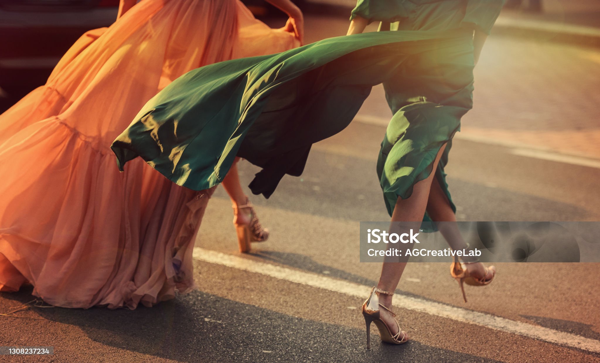 Two women in flowing peach and emerald gowns crossing a street at sunset